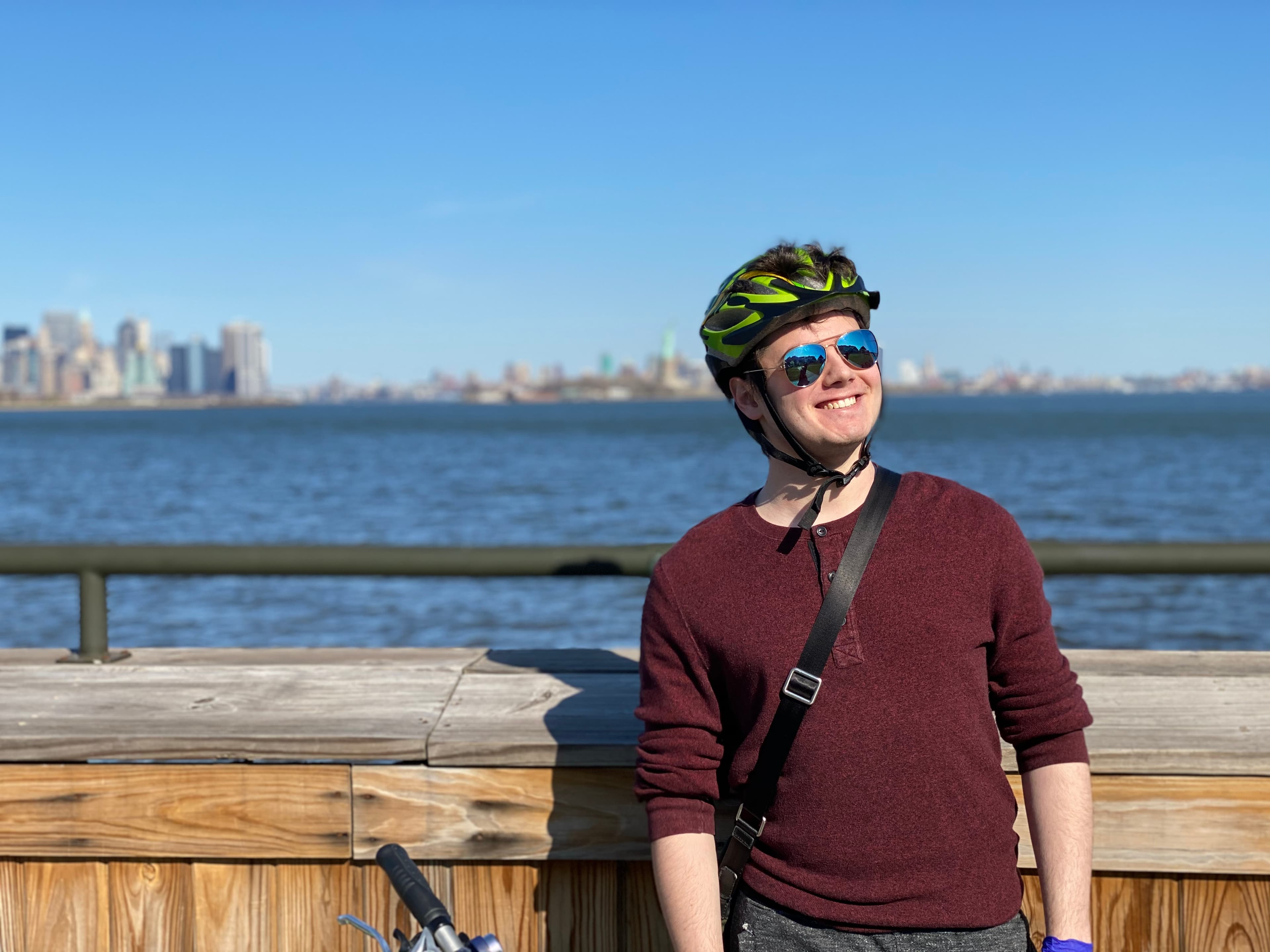 Hank Portney wearing a bike helmet, sunglasses, and a satchel in front of a railing overlooking Jersey City and the statue of liberty